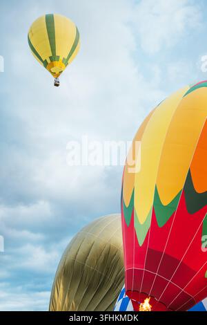 Ballons gonflables colorés vibrants en train de se gonfler avec une flamme active du brûleur au premier plan tandis qu'un ballon à rayures jaunes vole haut dans un ciel bleu nuageux pendant o Banque D'Images