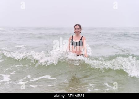 Jeune femme adulte aux cheveux bruns et à l'expression calme se tient dans une eau de mer peu profonde tandis que des vagues écumeuses se brisent autour d'un maillot de bain noir sous une lumière brumeuse douce Banque D'Images