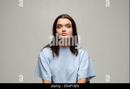 Jeune femme aux cheveux bruns et à l'expression neutre posant en t-shirt bleu sur un fond gris Banque D'Images