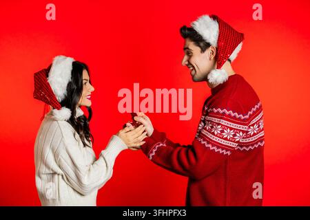 Jeune couple avec des chapeaux de Père Noël, l'homme fait sa demande en mariage à sa bien-aimée avec une bague sur un fond rouge en studio. Amour, h Banque D'Images