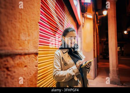 Jeune femme avec smartphone dans une rue nocturne de la ville. Dame debout seule sur un fond coloré, elle surfe sur Internet avec son mobile Banque D'Images