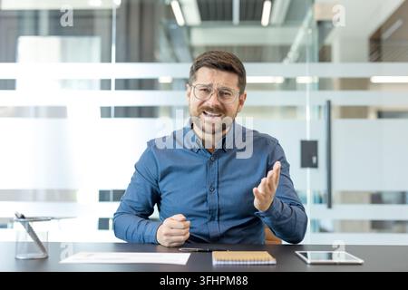 Communication en télétravail et stress des vidéoconférences, homme professionnel parlant lors d'une réunion en ligne dans un bureau moderne avec frustration. Banque D'Images