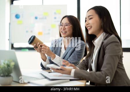 Deux femmes d'affaires collaborant dans Modern Office avec des ordinateurs portables et des ordinateurs portables, souriantes et engagées dans la discussion Banque D'Images