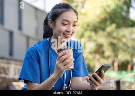 Travailleuse de santé asiatique en blouse bleue sourit tout en profitant d'un café et en utilisant son smartphone pendant une pause à l'extérieur. Banque D'Images