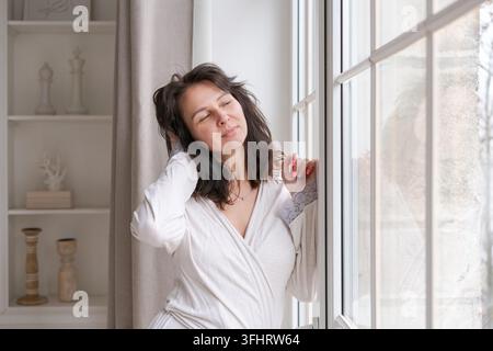Jeune femme riche et heureuse portant une robe de chambre la nuit devant une grande fenêtre dans la chambre et regardant Banque D'Images