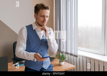 Jeune homme tenant un téléphone portable, communiquant et recevant un message texte qu'il lit Banque D'Images