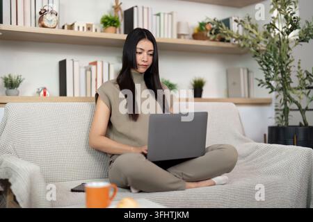 Une jeune femme est assise confortablement sur un canapé, concentrée sur son ordinateur portable, représentant l'essence du travail à distance et de la productivité. Banque D'Images