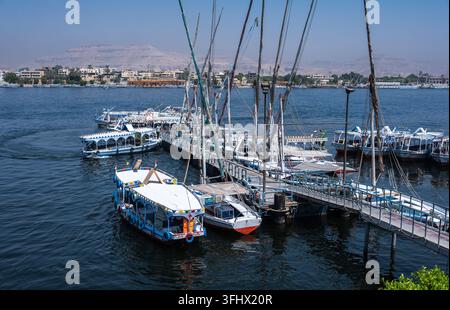 Louxor, Égypte - 27.07.2025. Les bateaux à voile traditionnels felouques et autres bateaux d'excursion sont amarrés à une jetée animée sur le Nil, prêts pour les touristes Banque D'Images
