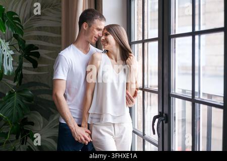Jeune couple souriant qui s'embrasse et se tient près d'une fenêtre, regardant Banque D'Images