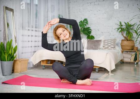 Femme âgée faisant de l'exercice assise en position du lotus. Femme mature active faisant des exercices d'étirement dans le salon à la maison. Banque D'Images