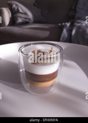 Boisson caféée en couches avec une épaisse mousse de lait et de la poudre de cacao dans un verre à double paroi transparent sur une table sous une lumière naturelle. Banque D'Images