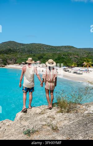 Un couple se tient la main tout en se tenant debout sur un bord rocheux, contemplant les eaux turquoises époustouflantes de Grote Knip Beach à Curaçao. Collines verdoyantes cadre th Banque D'Images