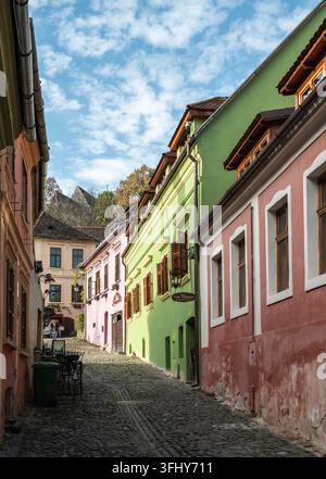 Sighisoara, Roumanie - 21.10.2025. Une vue charmante sur les maisons historiques roses, vertes et oranges vives bordant une étroite ruelle pavée montante en th Banque D'Images