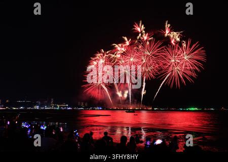 Des foules sur la plage de Pattaya regardent des feux d'artifice rouges éclatants explosant au-dessus de la baie océanique lors d'un festival nocturne en Thaïlande. Banque D'Images