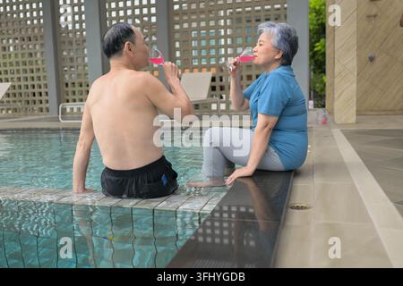 Couple senior assis près d'une piscine et toast des boissons ensemble, créant un moment de détente et de bonheur Banque D'Images