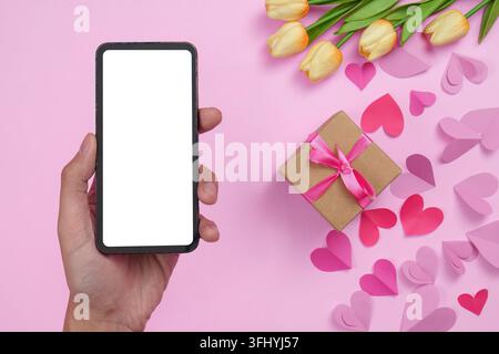 Un smartphone avec un écran vierge à côté d'une boîte cadeau avec un arc rose et des tulipes jaunes sur un fond rose, symbolisant la célébration et la communication. Banque D'Images