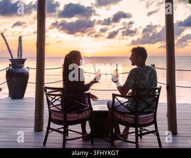 Deux personnes sont assises sur une terrasse en bois, sirotant des cocktails rafraîchissants en regardant le soleil se coucher sur la mer des Caraïbes à Sainte-Lucie. Le ciel est peint avec Banque D'Images