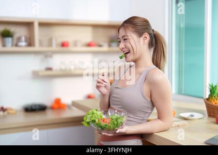 Une jeune femme en vêtements d'activité sourit tout en mangeant une salade fraîche dans une cuisine lumineuse et moderne, favorisant une alimentation saine et le bien-être. Banque D'Images