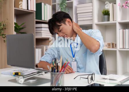 Homme asiatique souffrant de douleurs au cou et à l'épaule tout en travaillant au bureau Banque D'Images