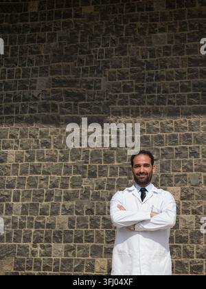 Médecin masculin du Moyen-Orient portant une blouse blanche et une cravate, debout les bras croisés, souriant avec assurance dans un cadre de clinique hospitalière. Professionnel m Banque D'Images