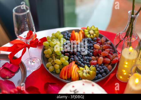 Plateau de fruits frais sur une table rouge romantique. Plateau de fruits frais avec raisins, myrtilles, kiwi et fruits coupés sur une table rouge romantique avec bougies Banque D'Images