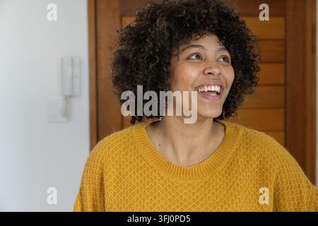 Femme afro-américaine en pull moutarde souriant regardant à la maison près de l'interphone et de la porte. Joie, bouclé, doré, tricot, chaleur, intérieur, entrywa Banque D'Images