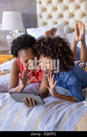 Amies diverses allongées sur un lit dans une chambre, tenant une tablette, faisant un signe de la main, sur une couverture à motifs. Confortable, chaleureux, intime, détendu, duo Banque D'Images