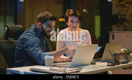 Deux collègues d'affaires multiethniques heureux avec un ordinateur portable et des documents de papier dans un bureau le soir. Homme d'affaires indien avec une femme d'affaires caucasienne Banque D'Images