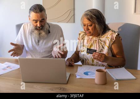 Couple senior examinant les finances sur ordinateur portable à la table à manger à la maison, tenant la carte de crédit, pointant. Partenaires, budgétisation, planification, paiement, intérieur, mug, Banque D'Images