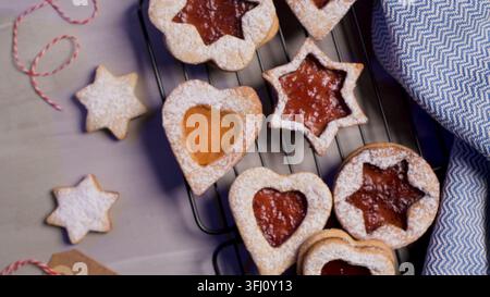 Vue de dessus de biscuits Linzer fraîchement cuits en formes d'étoile et de cœur, remplis de confiture rouge, saupoudrés de sucre glace sur une grille de refroidissement en fil métallique avec un b Banque D'Images
