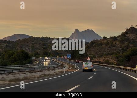 Benidorm, Espagne, 12 août 2026, autoroute à travers un paysage montagneux au coucher du soleil avec des véhicules sur la route et des pics dramatiques en arrière-plan en sp Banque D'Images