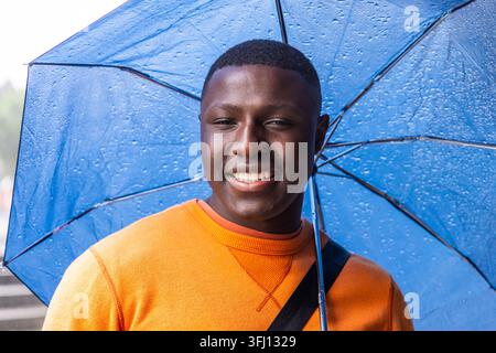 Homme afro-américain debout sous un parapluie bleu dans une passerelle urbaine pluvieuse, portant un sweat-shirt orange. Pluie, extérieur, parasol, vibrant, décontracté, souriez, Banque D'Images