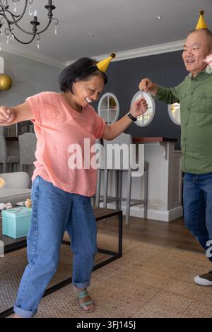 Couple de femme âgée et homme mûr dansant dans le salon avec des chapeaux de fête et des cupcakes. Célébration, maison, festif, cosy, bougie Banque D'Images