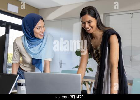 Diverses collègues féminines se penchant sur un ordinateur portable argenté dans la salle de réunion, portant le hijab et la tunique. Collaboration, travail d'équipe, collègues, espace de travail, minimali Banque D'Images