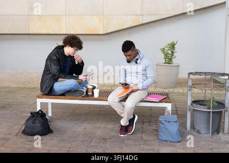 Divers étudiants masculins assis sur un banc à lattes en bois à la place du campus en utilisant des smartphones, des ordinateurs portables. Duo, éducation, photoréaliste, stock, extérieur, ur Banque D'Images