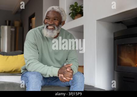Homme afro-américain senior assis sur un pouf souriant et regardant laissé par le poêle à bois. Personnes âgées, gentleman, assis, confortable, chaleur, maison, plante Banque D'Images