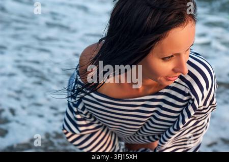 Jeune femme caucasienne brune en chemise rayée style marin avec les cheveux mouillés, souriant au bord de la mer vue de dessus Banque D'Images