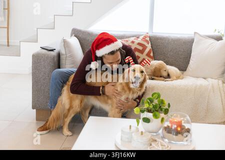 Homme adulte moyen portant un chapeau de père Noël rouge embrassant doré avec bandeau en bois de renne sur le canapé. Chien, confortable, vacances, festif, salon, décor, env Banque D'Images