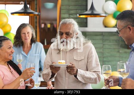 Famille diversifiée et amis se réunissant autour du comptoir dans la cuisine, homme senior tenant la bougie cupcake. Célébration, ballons, maison, intime, toast, wi Banque D'Images