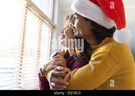 Couple diversifié embrassant à la maison par stores de fenêtre homme portant chapeau de Père Noël pulls smartwatch Sunlit. Romantique, intime, confortable, vacances, lumière du jour, câlin, Banque D'Images