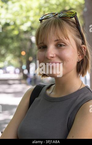 Femme debout et souriant légèrement sur le trottoir ensoleillé portant des lunettes de soleil sac à dos et arceau de nez. Portrait, extérieur, urbain, bokeh, ensoleillé, décontracté, blon Banque D'Images