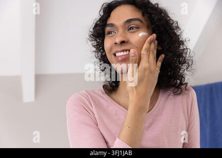 Personne non binaire appliquant la crème blanche à la joue à la vanité, portant le dessus rose, les bagues et le collier. Hydratant, soin de la peau, soin de soi, cheveux frisés, lumière douce Banque D'Images
