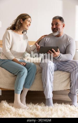 Couple assis sur un lit tenant une tablette, homme mûr en pyjama rayé, femme en pull crème. Confortable, chaud, intime, détendu, inte Banque D'Images