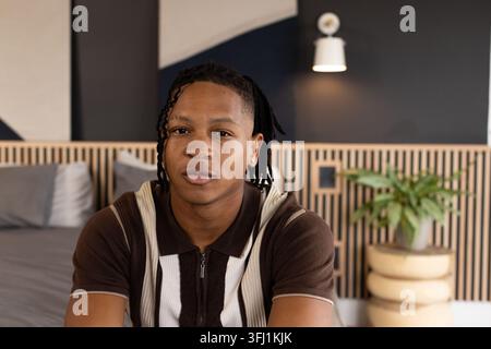 Homme afro-américain dans la vingtaine assis sur un lit avec tête de lit en bois portant un haut en tricot marron Banque D'Images
