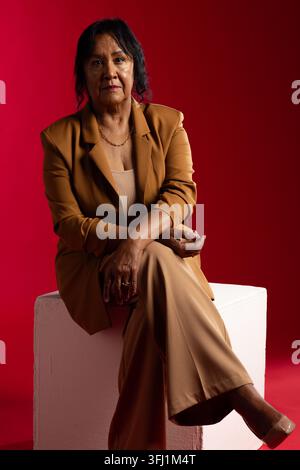 Sénior afro-américaine assise sur un cube blanc dans un studio portant un costume beige et un collier chaîne Banque D'Images