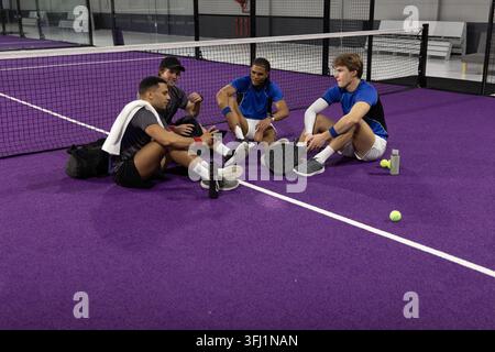 Quatre athlètes masculins adultes en vêtements de sport assis sur un court violet prenant une pause, tenant des pagaies, de l'eau. Pickleball, coéquipiers, casual, style de vie, sports, Banque D'Images