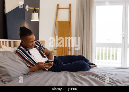 Homme afro-américain d'une vingtaine d'années allongé sur un lit, utilisant un smartphone et portant des écouteurs dans la chambre Banque D'Images