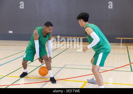 Divers joueurs masculins portant des maillots verts pratiquant le dribble de basket-ball orange sur le terrain. Coéquipiers, défense, travail d'équipe, bois dur, gymnase, banc, Banque D'Images
