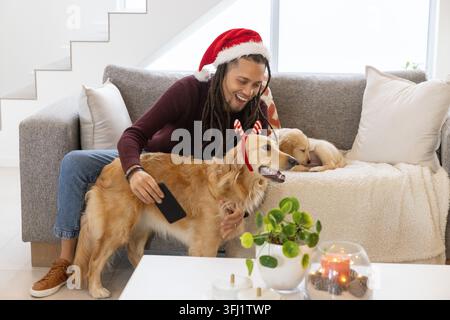 Mi-30s homme portant chapeau de Père Noël se penchant souriant tenant téléphone caressant Golden retriever sur canapé gris. Chien, chiot, bandeau, bois, confortable, festif, brick Banque D'Images