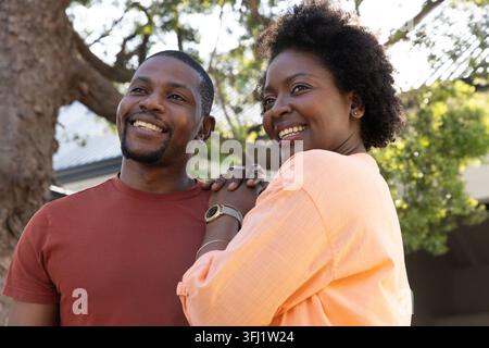 Couple afro-américain souriant, debout près d'une maison sous un arbre, montrant une montre et un bracelet. Ensoleillé, intime, romance, harmonie Banque D'Images